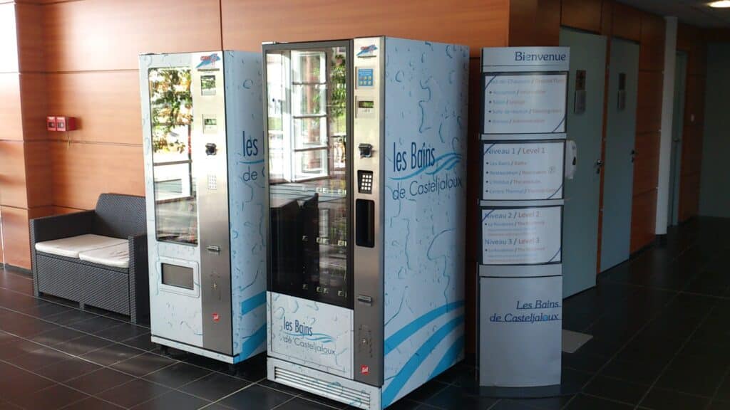 Deux distributeurs automatiques personnalisés aux couleurs des Bains de Casteljaloux, installés dans un espace intérieur moderne avec des panneaux d'information et un coin salon.